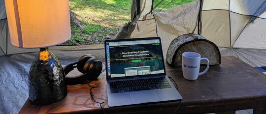 Outdoor workspace setup with a lamp, headphones, laptop and Checkfront mug on a table inside a tent