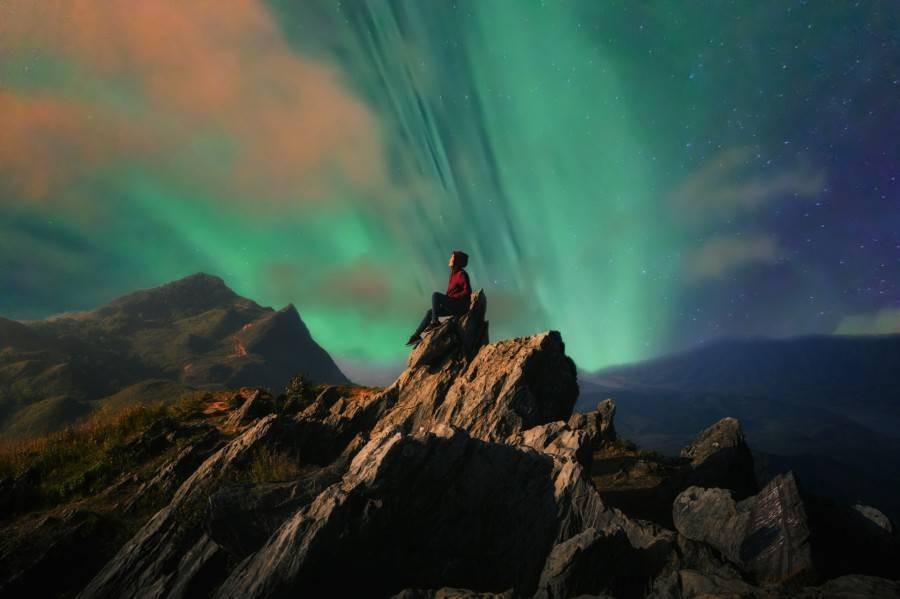 Woman in red coat sitting at the peak of a mountain looking at the northern lights above
