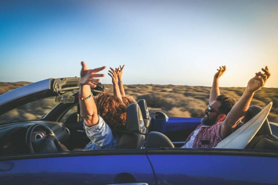 Group of travelers riding in convertible in the desert with their hands up in celebration