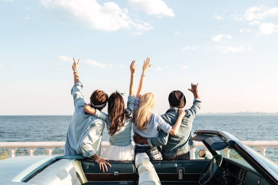 Back view of happy young friends standing with raised hands sitting the car looking at ocean