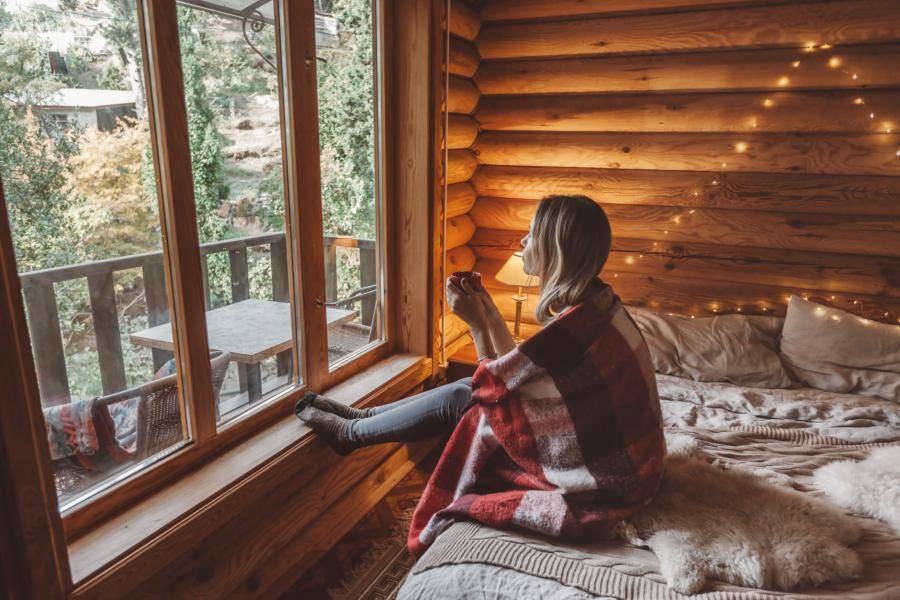 A person looking very cozy as they sip coffee and stare out a huge window into the forest