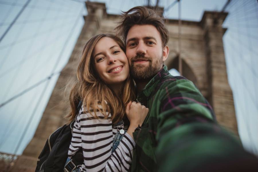Young couple taking a selfie standing in front of Brooklyn Bridge on New York City tour.