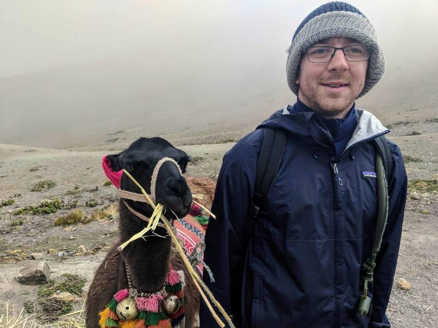 Male solo traveler posing with alpaca while hiking up Rainbow Mountains in Peru