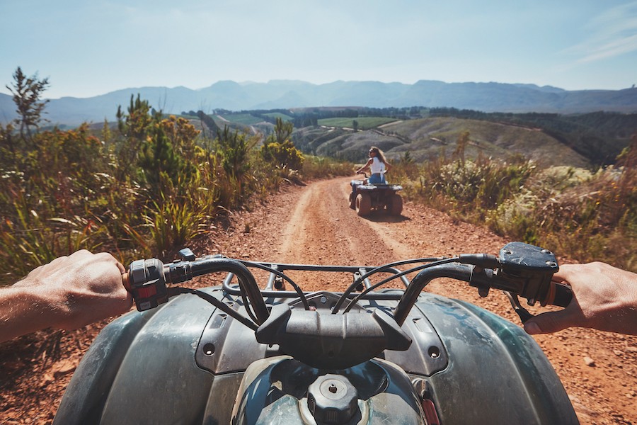 View from a quad bike in nature with a woman in front driving off road on an all terrain vehicle. POV of a quad biker following another ATV on a trail